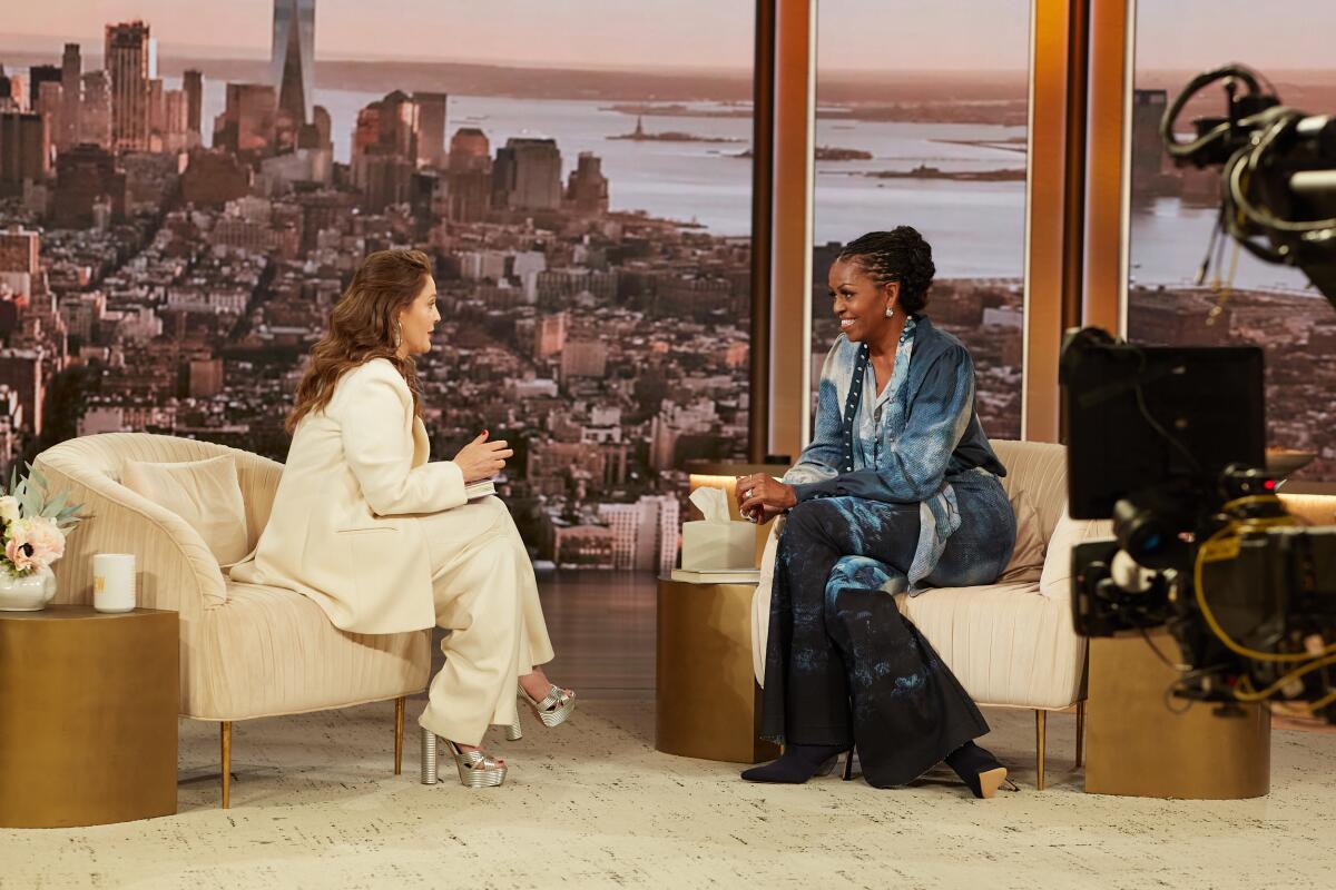 Drew Barrymore and Michelle Obama talk facing each other while sitting in chairs on a TV set.