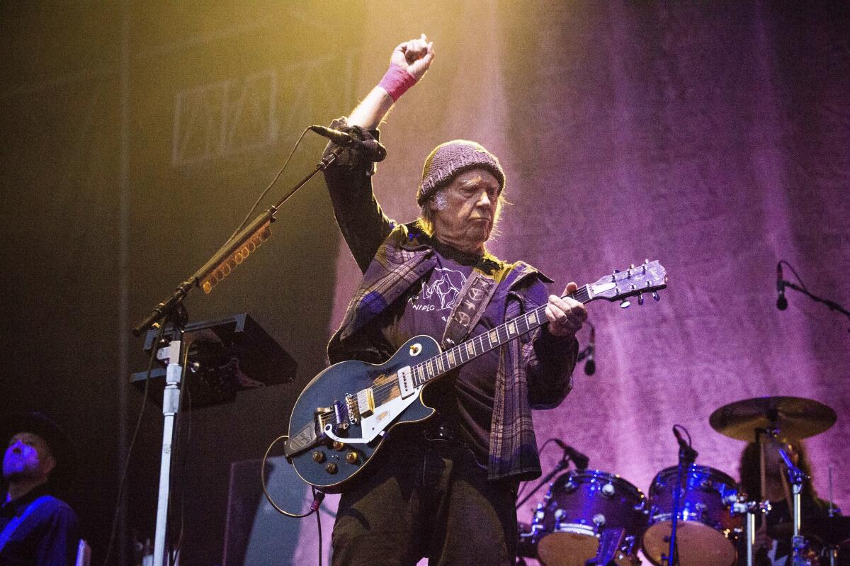 Neil Young holding a guitar in concert in a dark jacket, dark pants and a knit beanie with his right arm raised