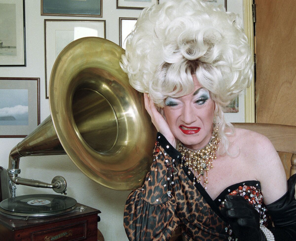 Paul O'Grady as Lily Savage, with a big blond bouffant, wearing a one-shoulder gown and standing next to a gramophone.