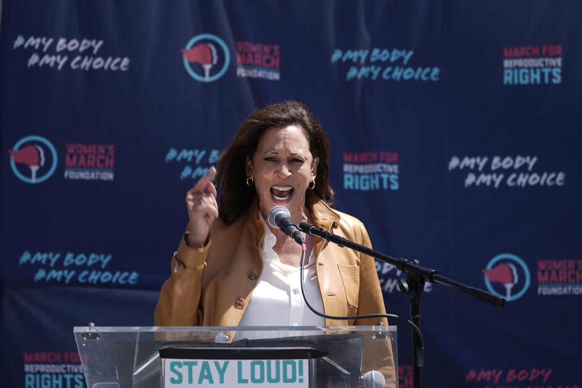 Vice President Kamala Harris speaking into a microphone in front of a banner with messages including "My body/My choice."