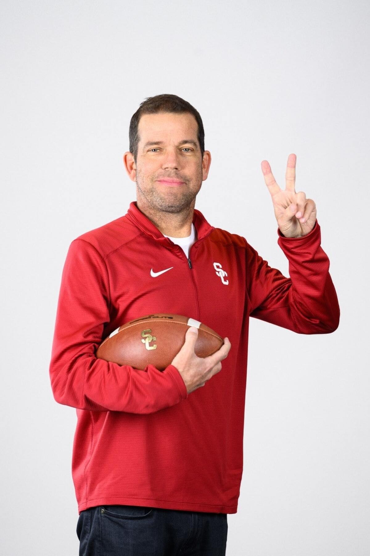 USC coach Dave Nichol holds a football in one hand while holding up two fingers in "V" shape.  