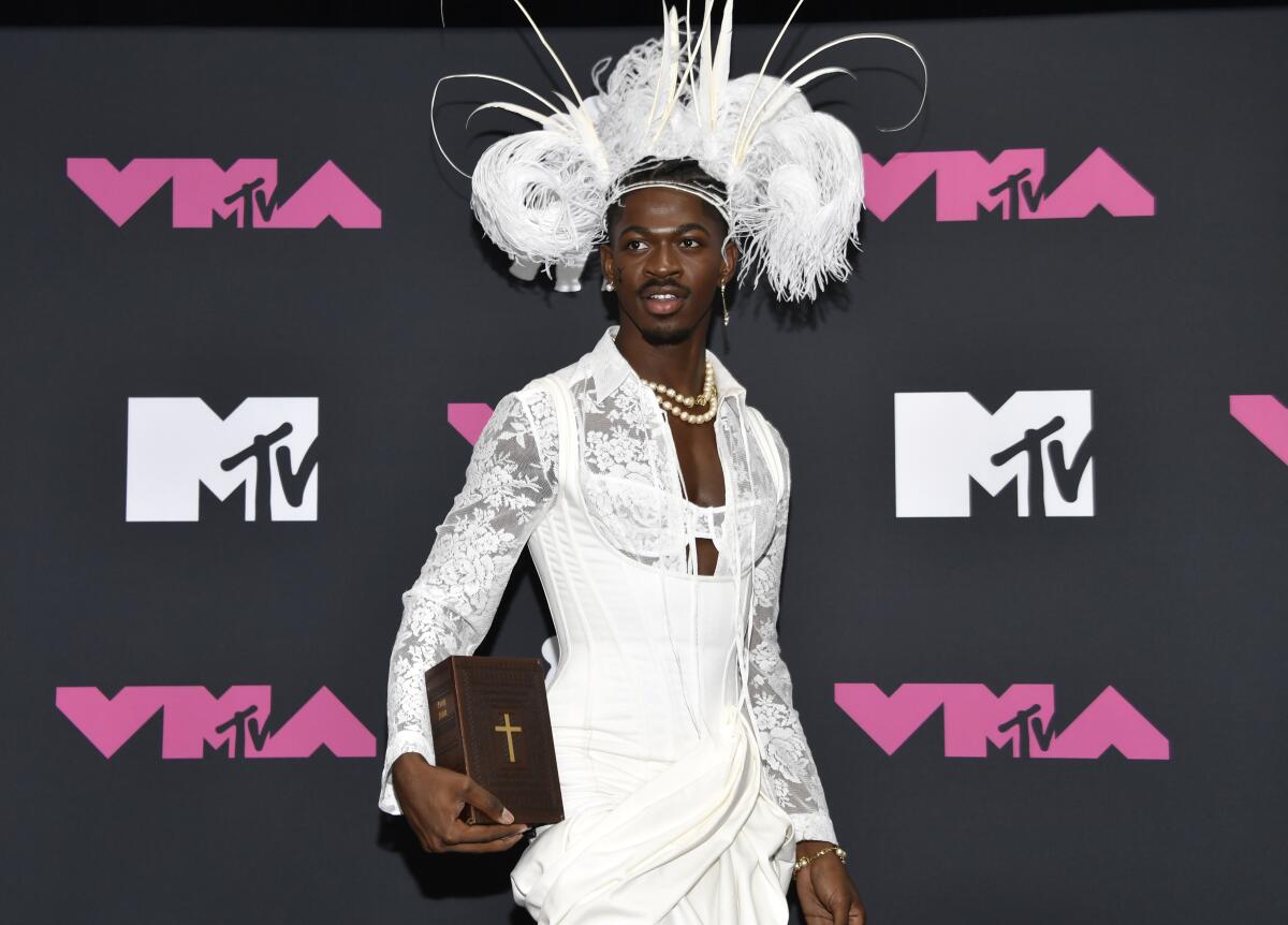 Lil Nas X poses in a white corset and feathered headdress holding a Bible next to his right hip