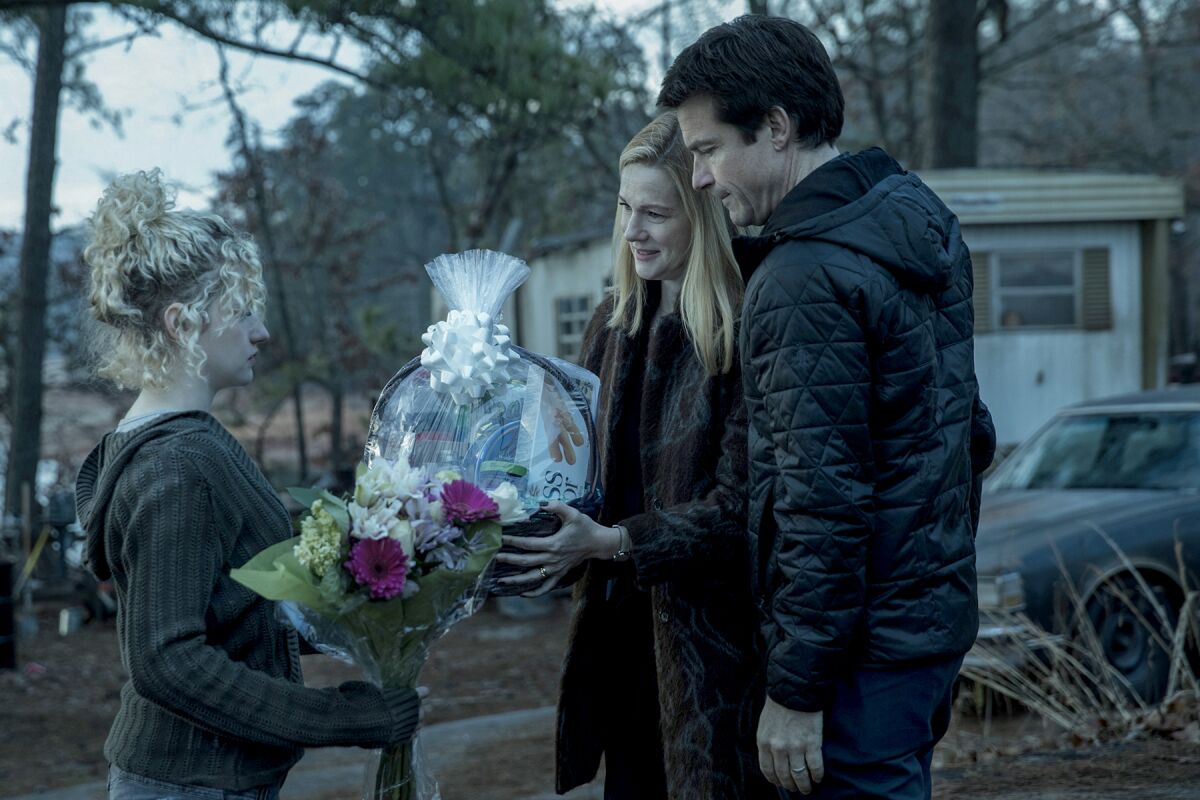 A woman holding flowers stands in front of a man and a woman holding a gift basket