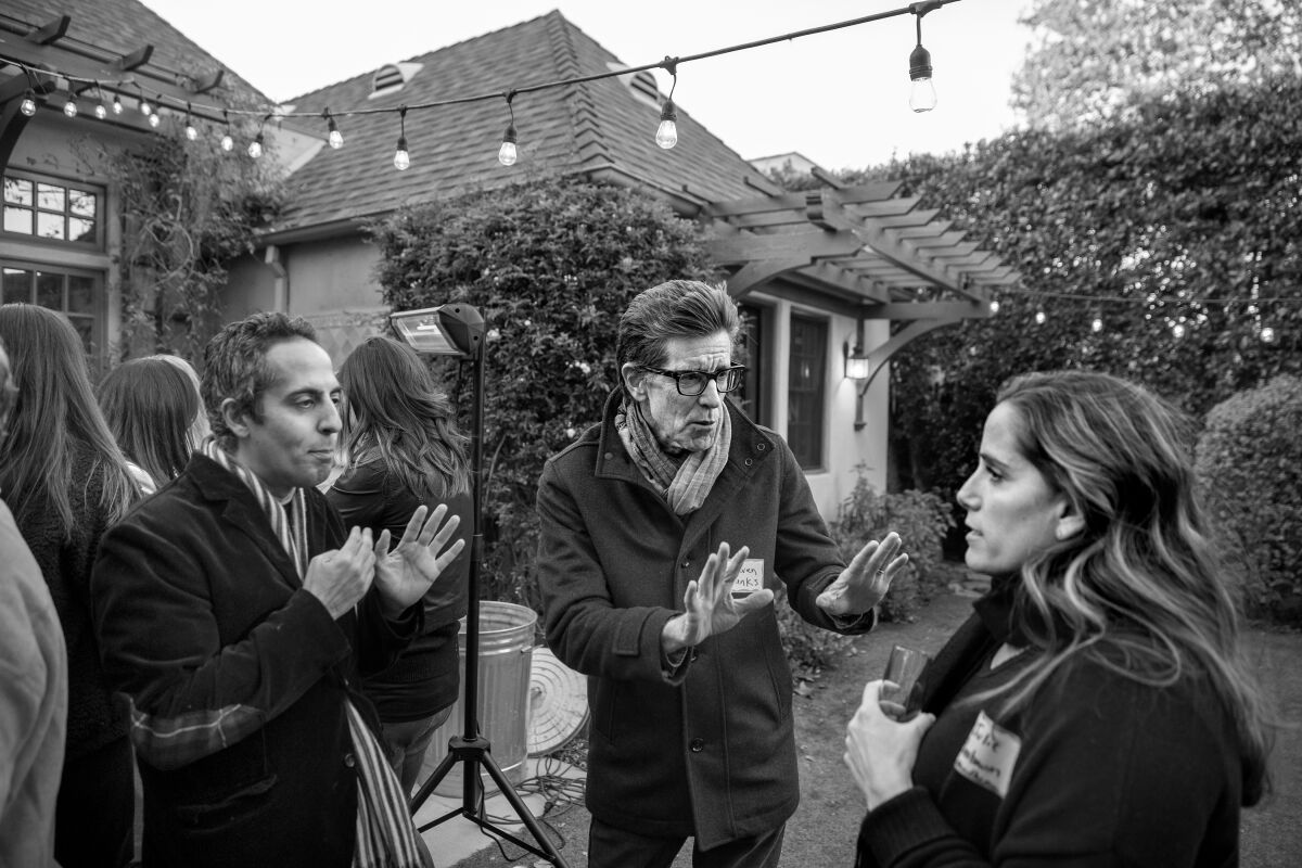 Two men talk to a woman at a gathering outdoors in a backyard.