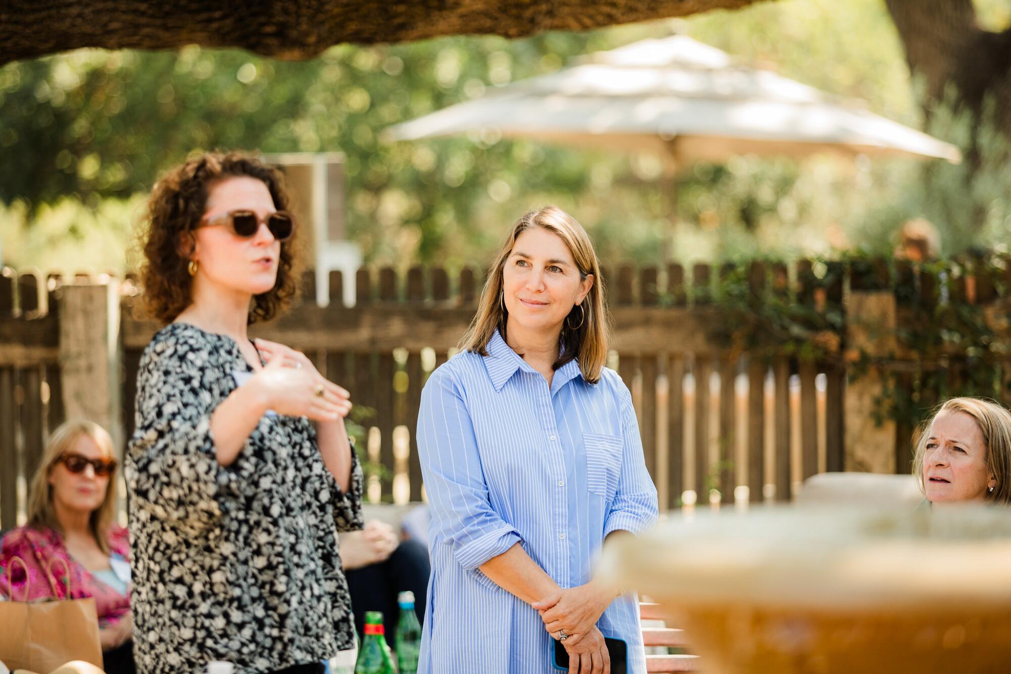 Two women stand outside, addressing people seated at tables around them.