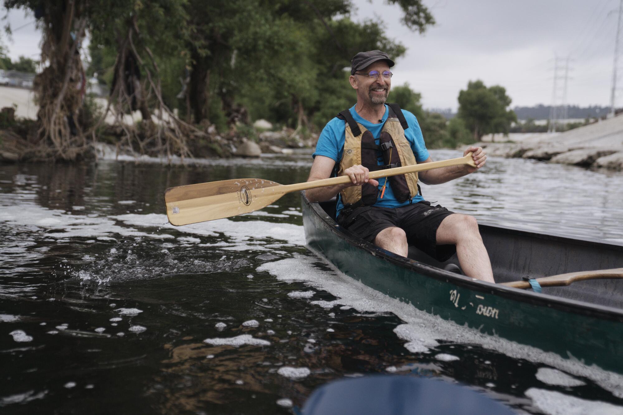 A man in a canoe grins as he holds a paddle above the waters of a river.
