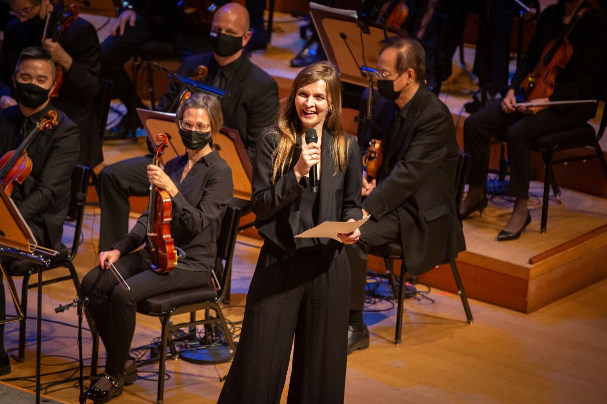 A woman in a black suit, microphone in hand, stands in front of black-clad orchestra members.