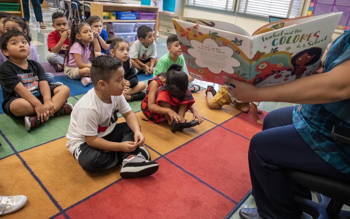 Children sit cross-legged on a multicolored rug as a teacher reads a picture book from a chair in a kindergarten classroom