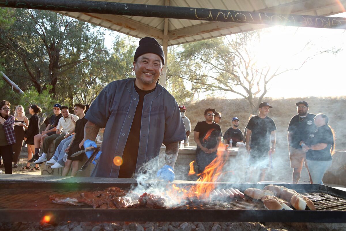A smiling man stands before a large flaming grill