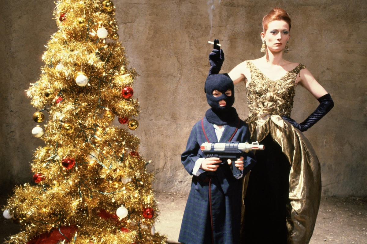 A boy in a full face mask holding a weapon and a glamorous woman pose in front of a Christmas tree.