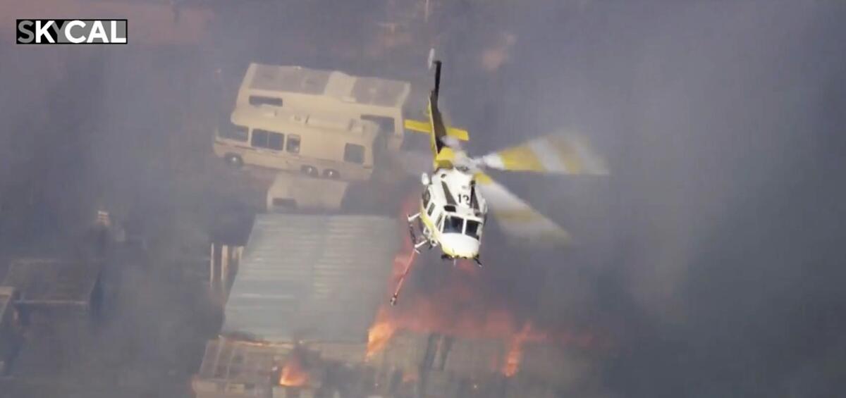 A helicopter flies in smoky air over a small, metal-roofed building on fire. Nearby is an RV.