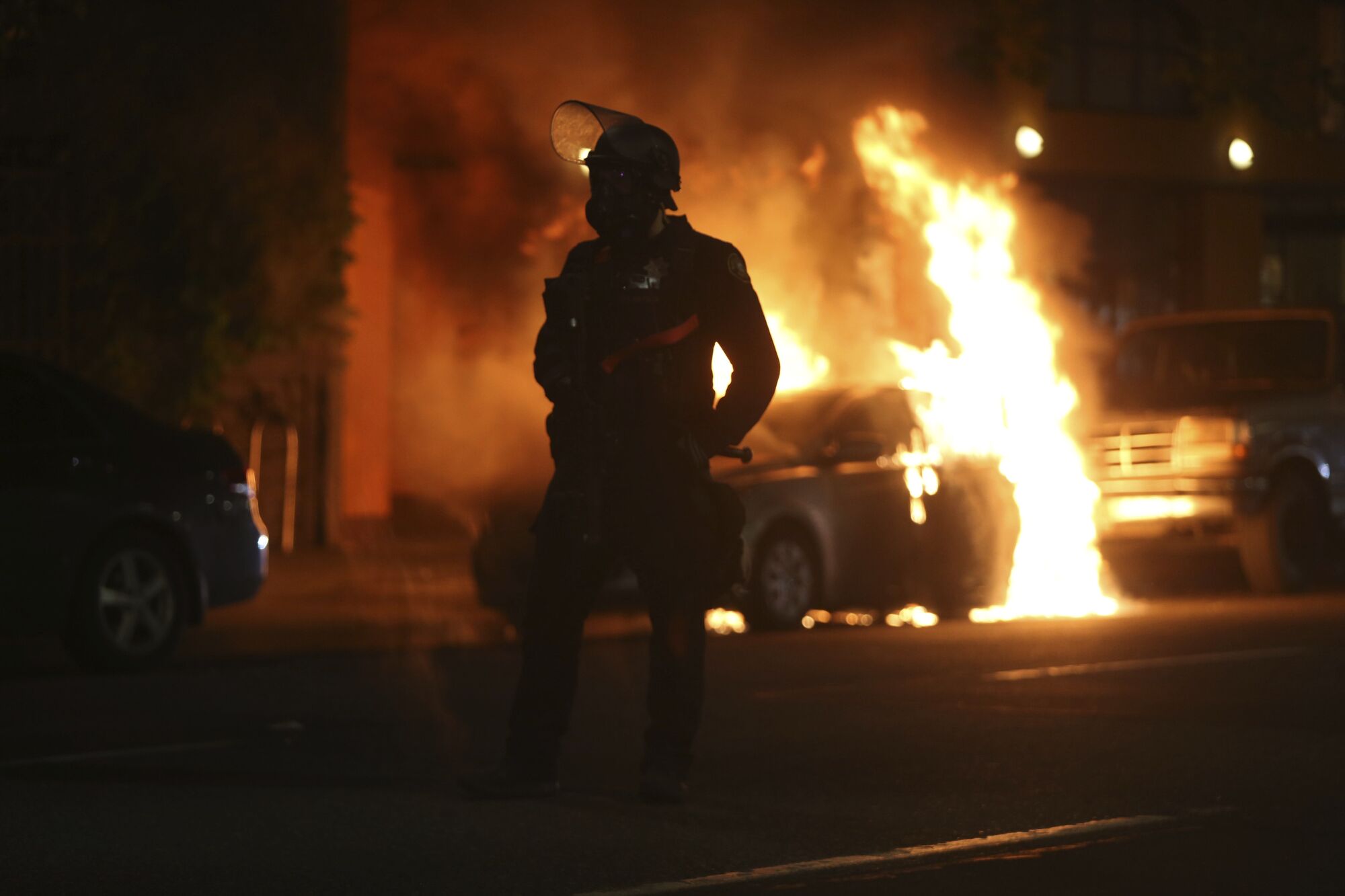 A police officer is silhouetted near a burning car.