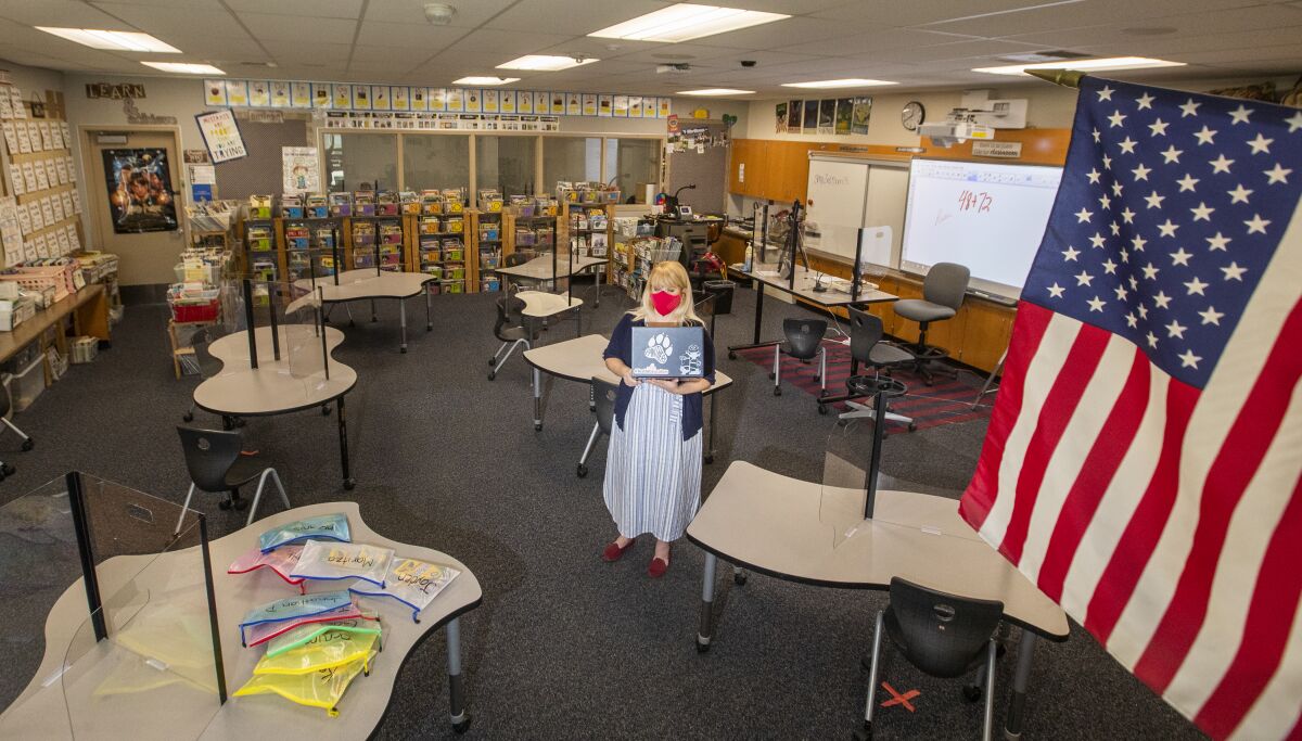 A teacher wearing a red face mask stands in her empty classroom.