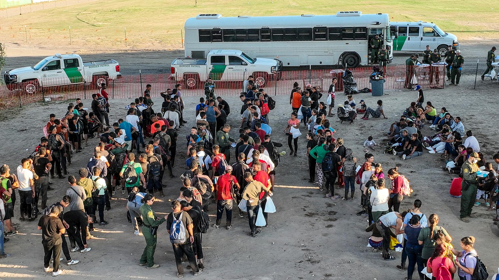 An aerial view of migrants forming lines in a barren lot beside several vehicles