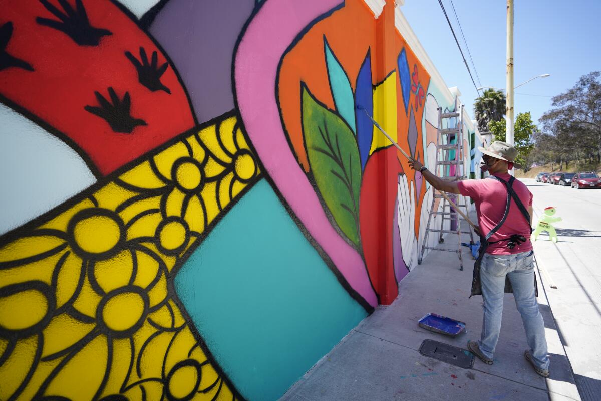 A man in red shirt and tan hat paints a colorful mural on the side of a medical clinic