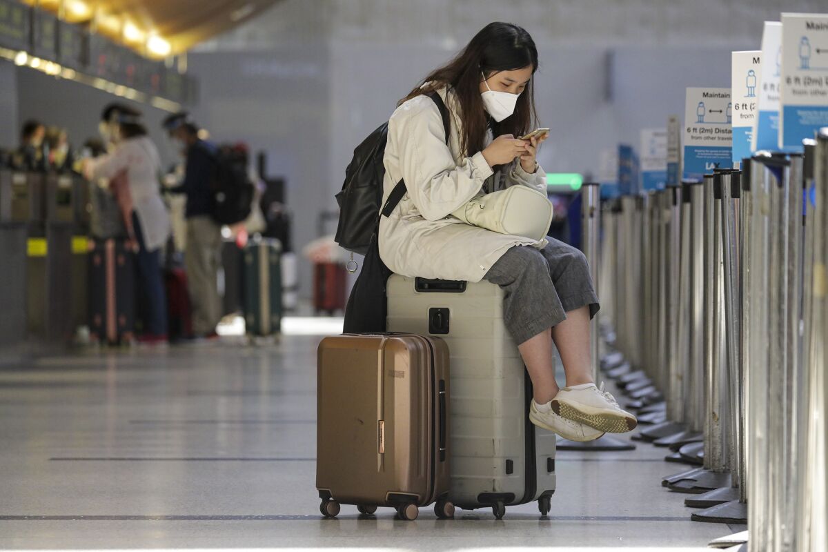 A woman in a mask sits on her luggage while looking at her phone