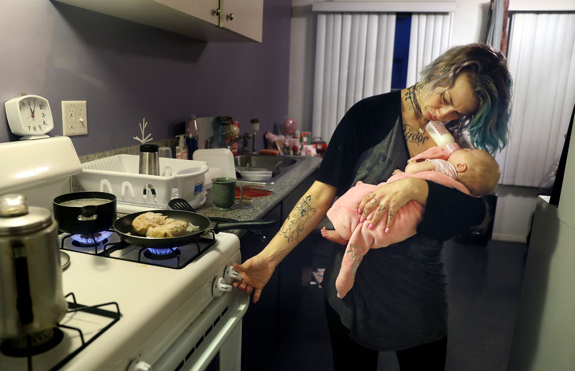 A woman carrying a baby holds a baby bottle against her chin while turning the nob on a stove to cook dinner