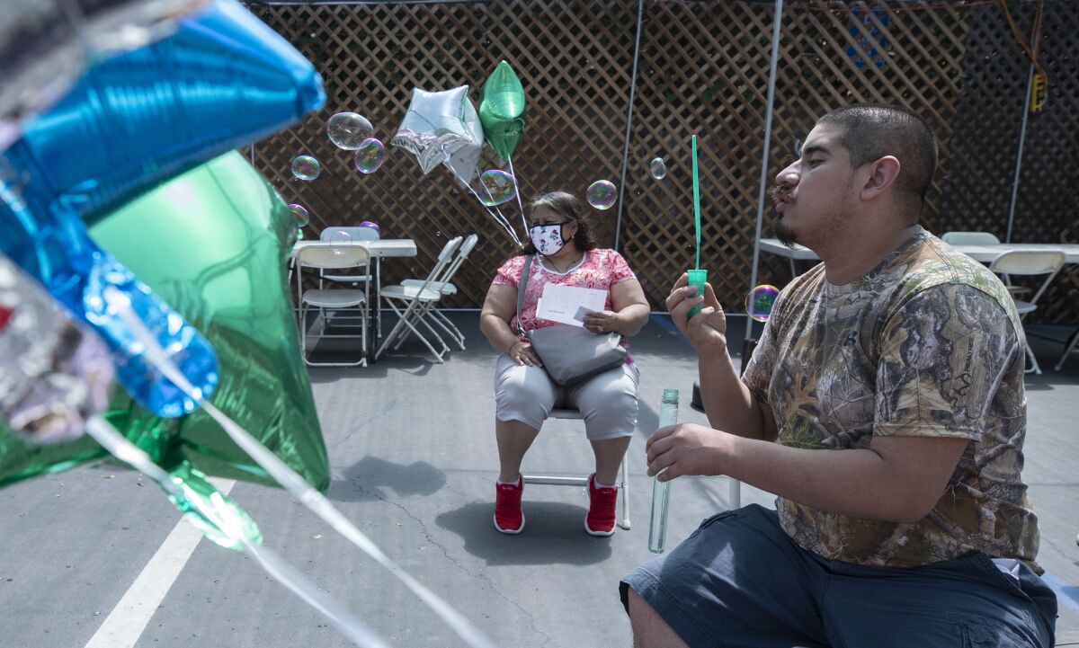 A man sits in a chair blowing bubbles outside. Green, silver and blue balloons are also in view. A woman is also seated.