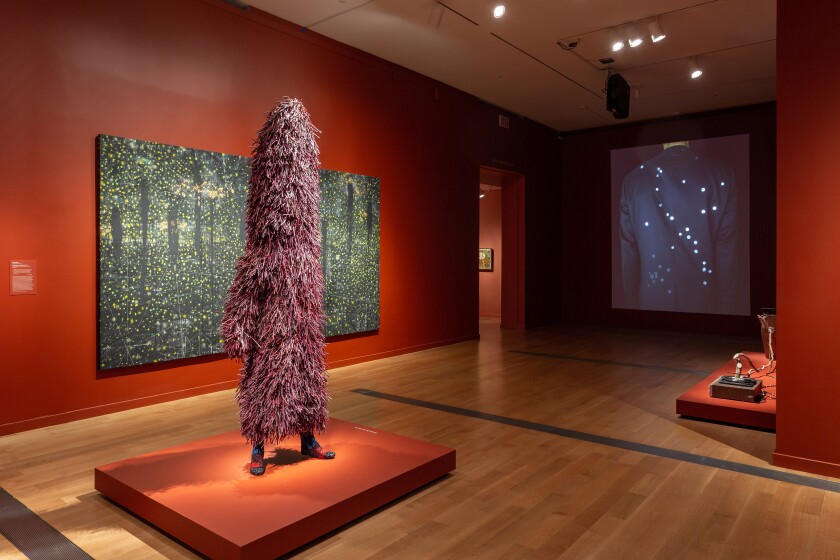 A view of gallery with red walls features a human figure covered in a suit made of twigs before a starry painting