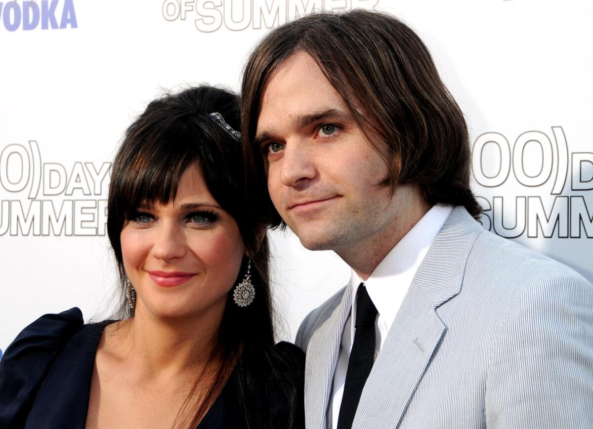 Zooey Deschanel, in a dress, and Ben Gibbard, in a light gray suit, smile for photos.
