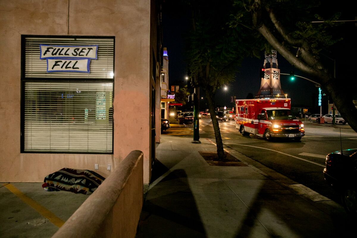 A homeless person sleeps on the ground as an ambulance passes by outside a shuttered nail salon.