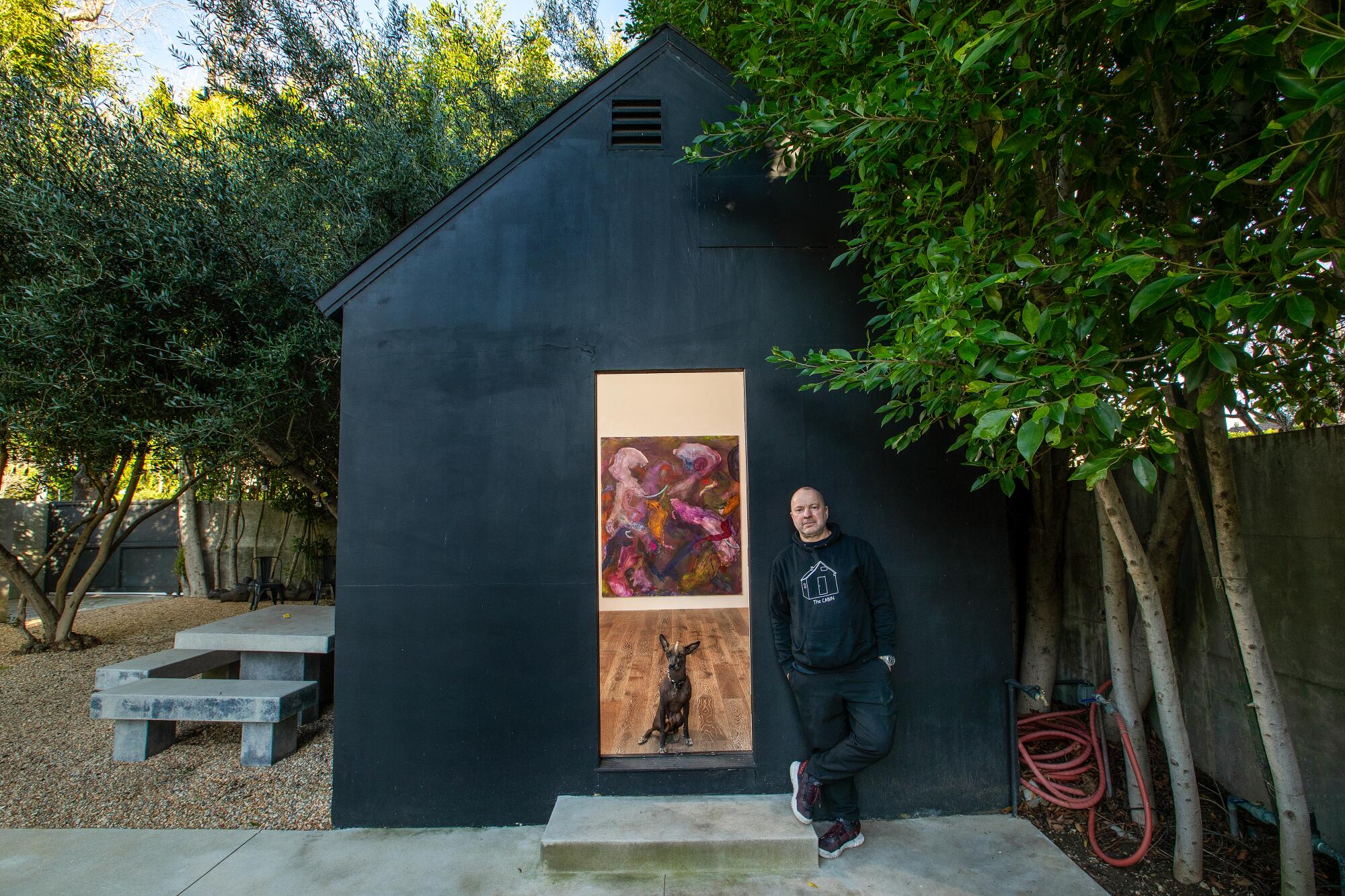 A man stands outside a small building, painted black, with a dog and paintings visible through its open door
