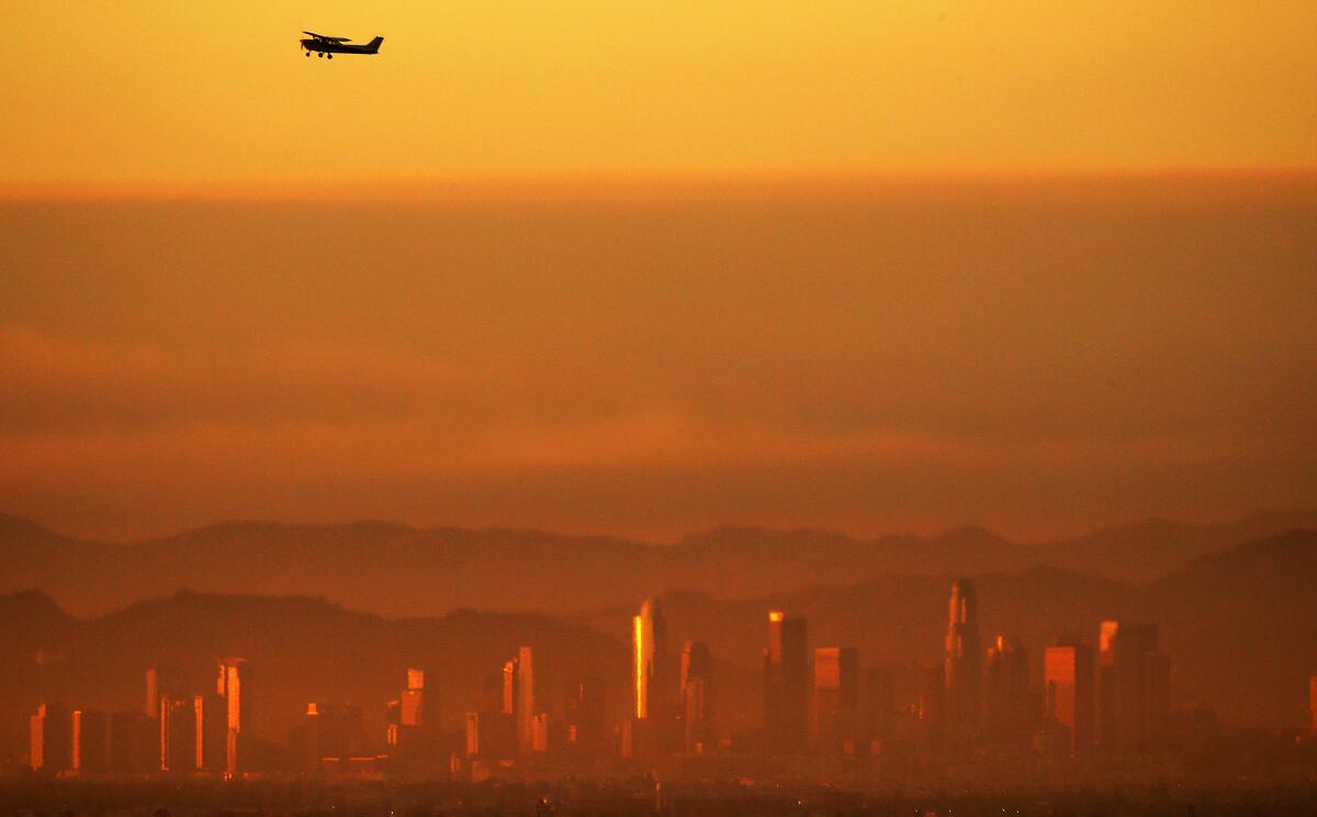 The downtown Los Angeles skyline shimmers in the smog as a plane taking off from Long Beach Airport climbs out of the haze.