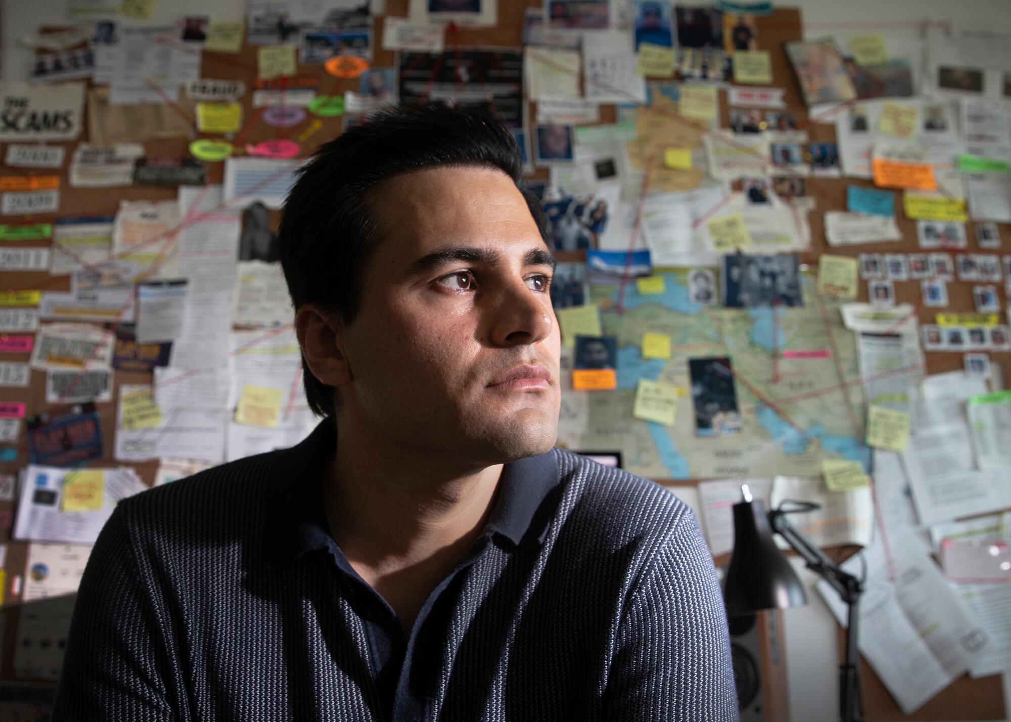 A man with dark hair, framed head-and-shoulders, sits in front of a wall with news clippings and pictures, looking to right