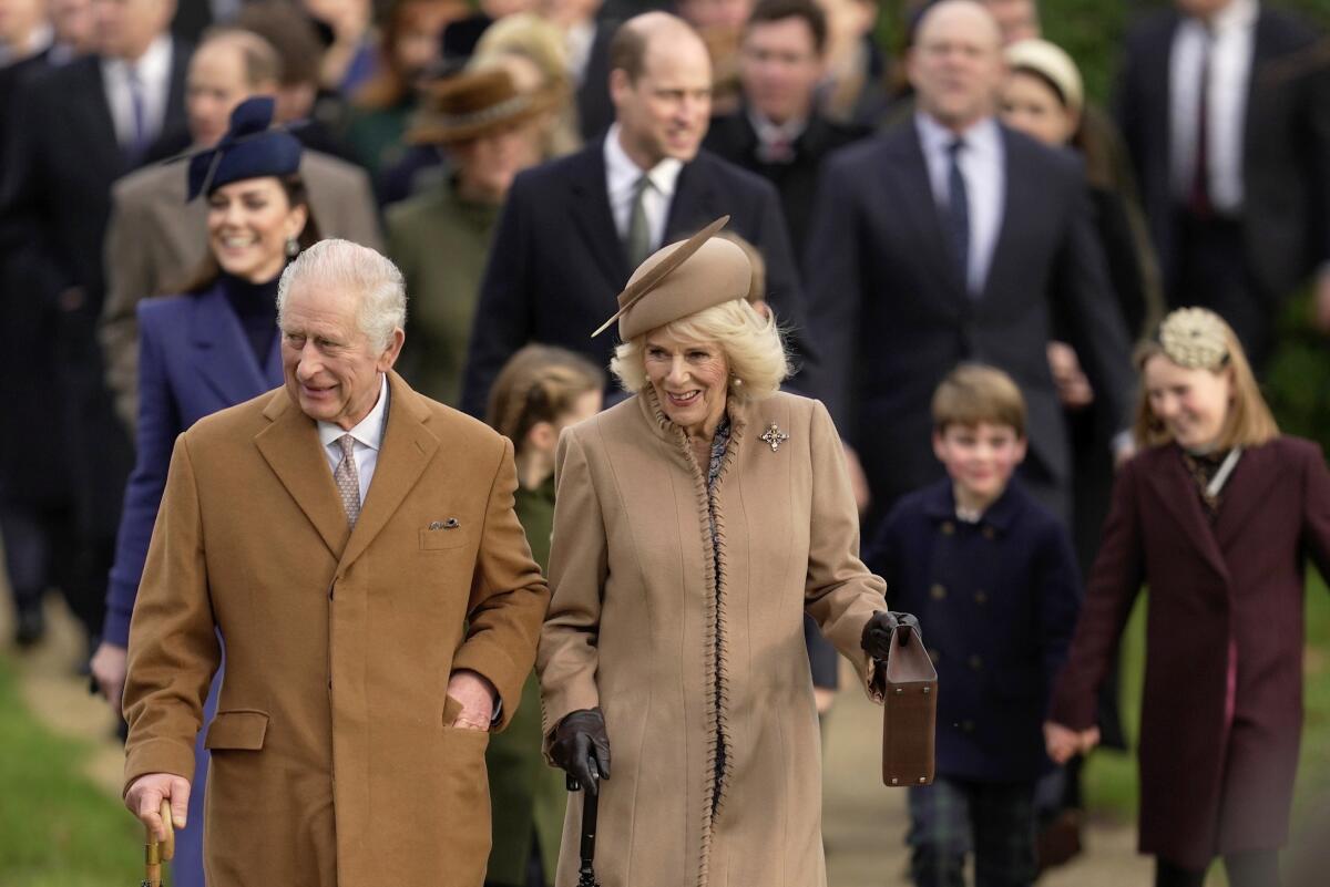 Britain's King Charles III and Queen Camilla walk outdoors wearing coats followed by other members of the royal family.