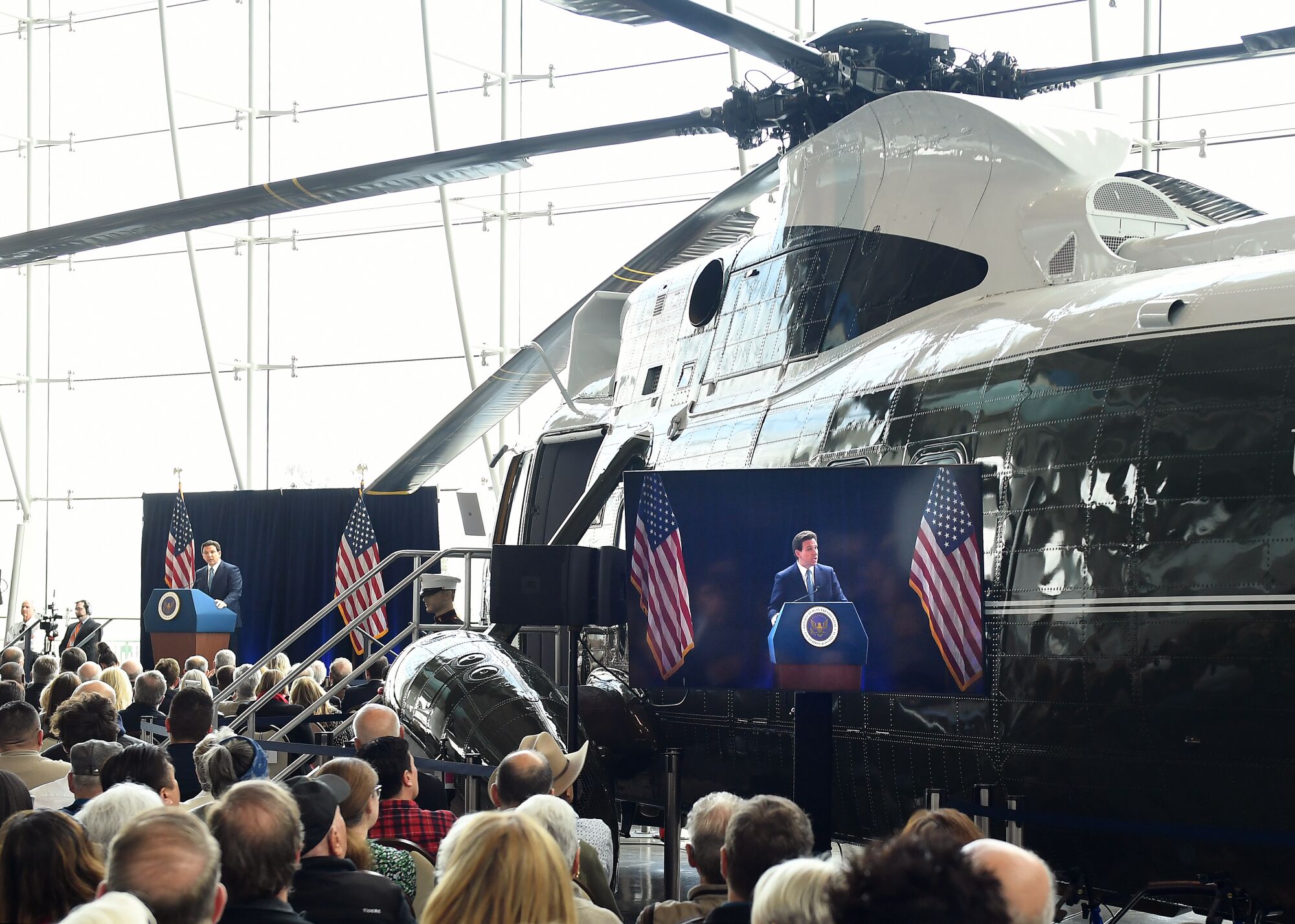 a man in a suit and tie speaks from a podium in front of a retired presidential helicopter. 