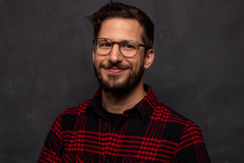 Andy Samberg smiling in glasses and a red and black plaid shirt in front of a dark background