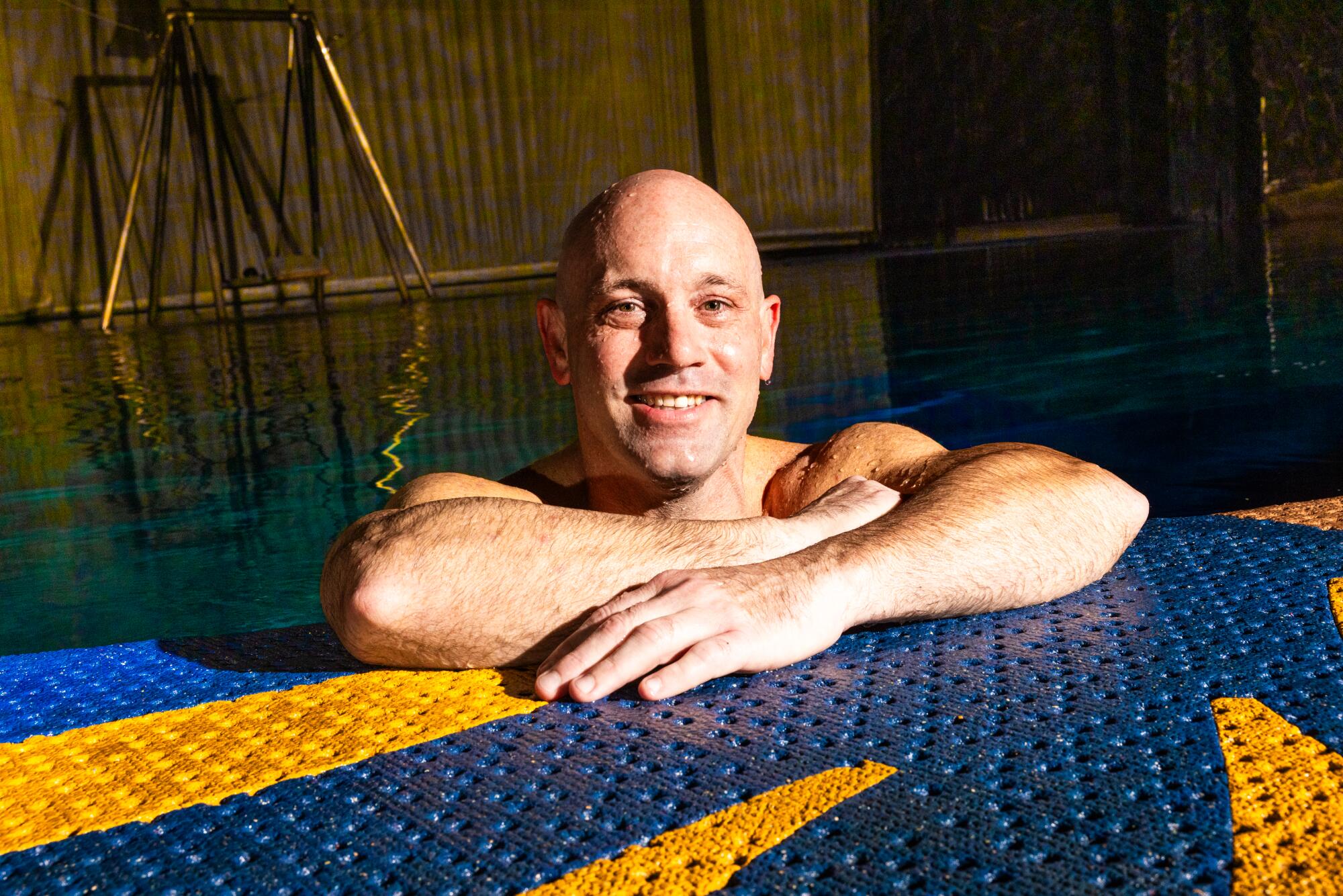 A bald, muscular man submerged chest down in the water smiles at the camera. 
