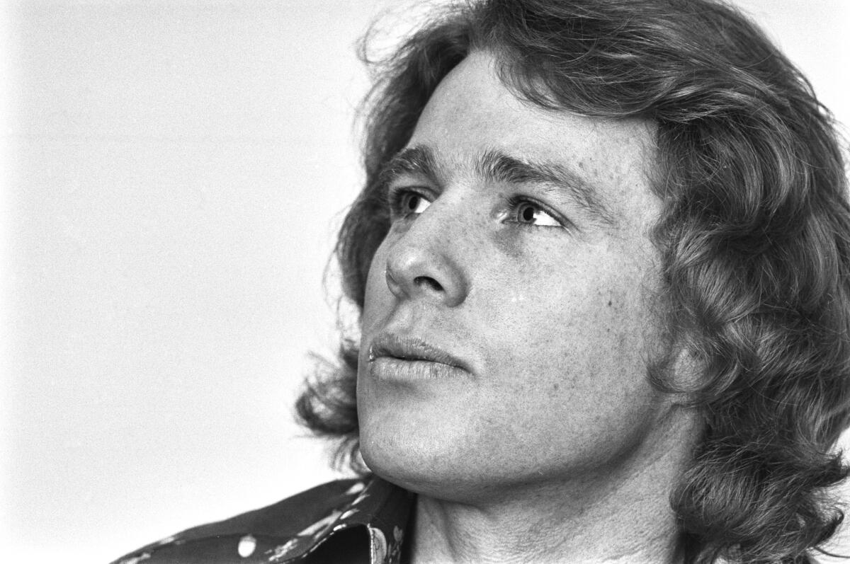 A youthful Ryan O'Neal, with shoulder-length curly hair, stares to the side with a slight smile.