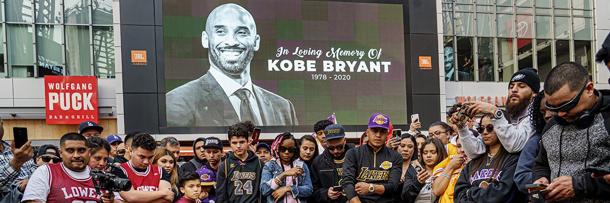 A crowd of fans stands outside of Staples Center in front of a screen with the words "In loving memory of Kobe Bryant 1978-2020" and Bryant's photo.