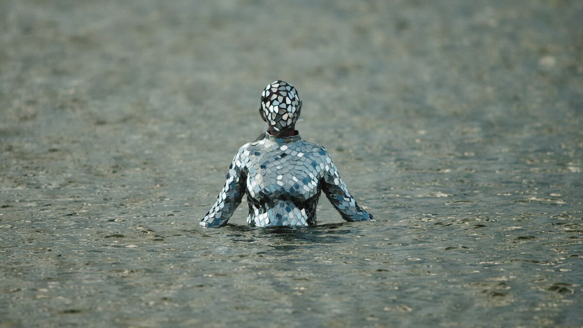 A mysterious female figure, her entire body and head encased in a silver bodysuit, emerges from a body of water.