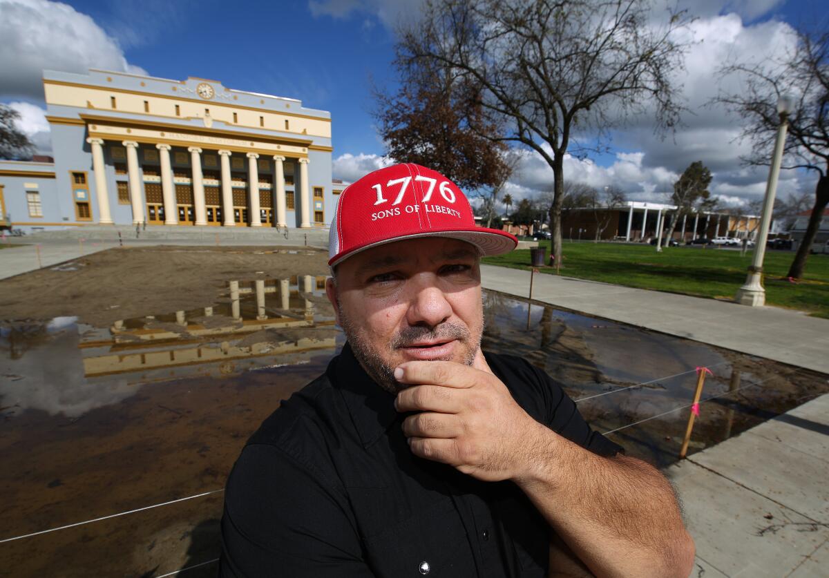 A man in a red hat that says 1776 stands outside near buildings while holding his chin.