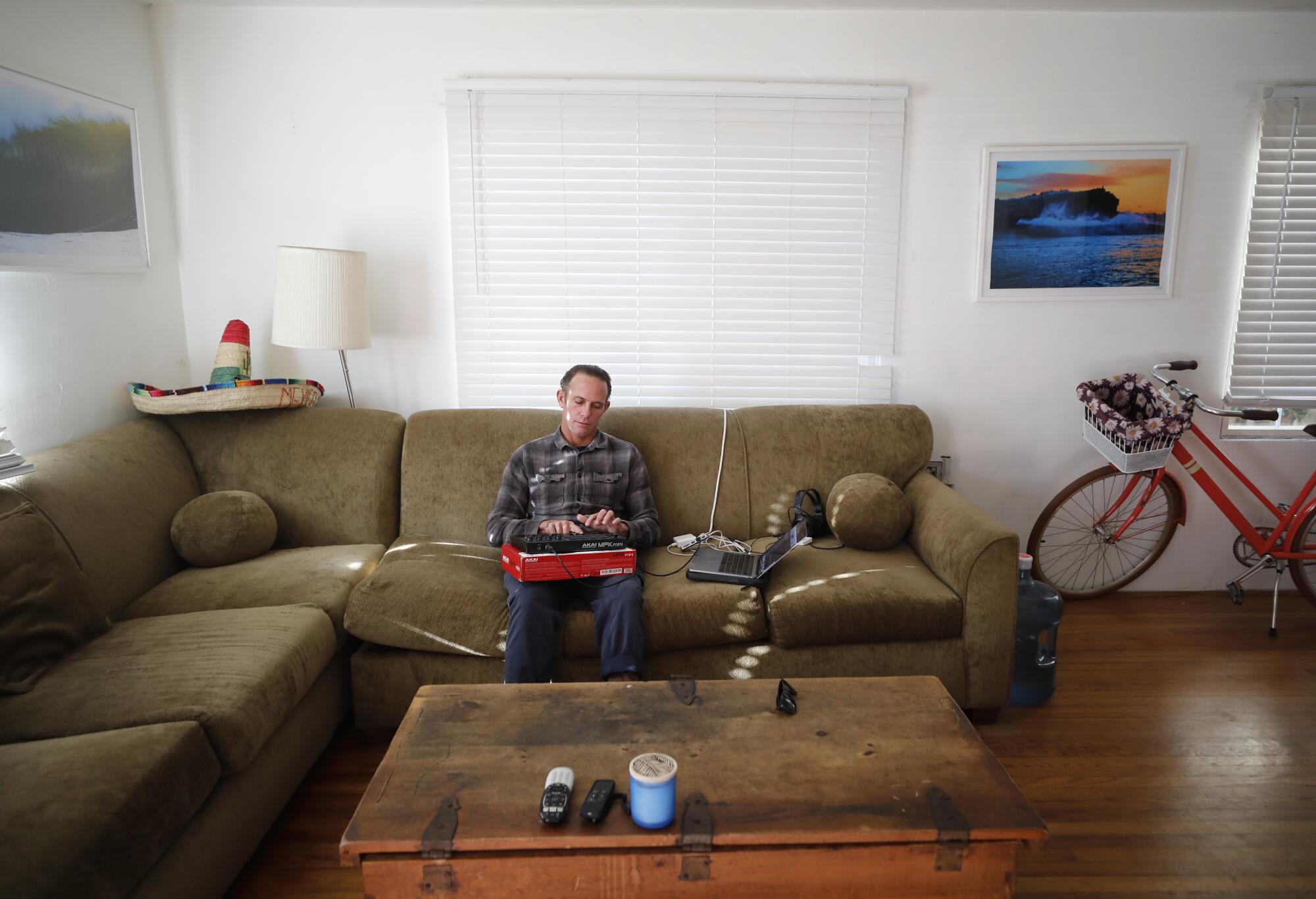A man sits on a couch, playing a keyboard plugged into a laptop