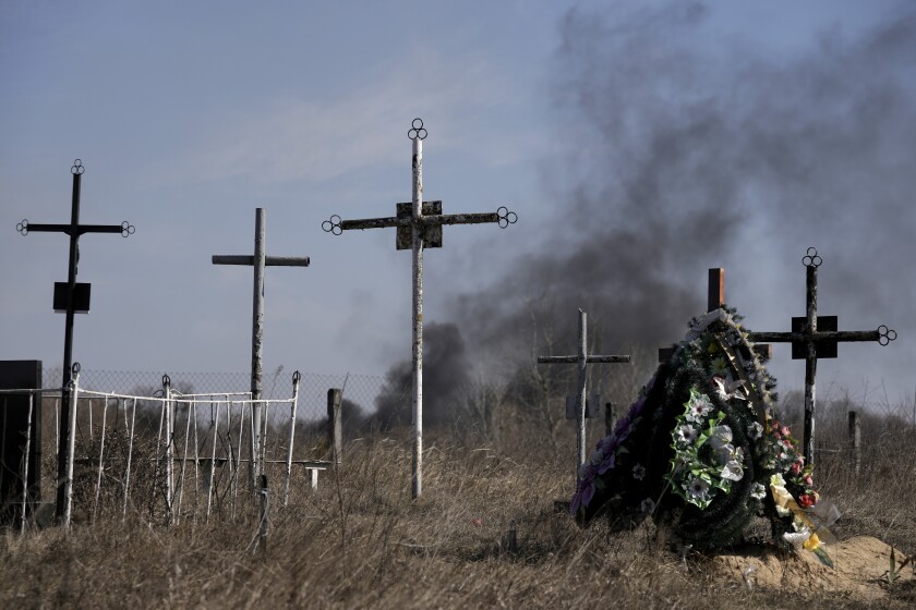 A smoke from shelling rises as a wreath of flowers is placed at a cemetery in Vasylkiv south west of Kyiv, Ukraine, Saturday, March 12, 2022. Russian forces appeared to make progress from northeast Ukraine in their slow fight to reach the capital, Kyiv, while tanks and artillery pounded places already under siege. (AP Photo/Vadim Ghirda)