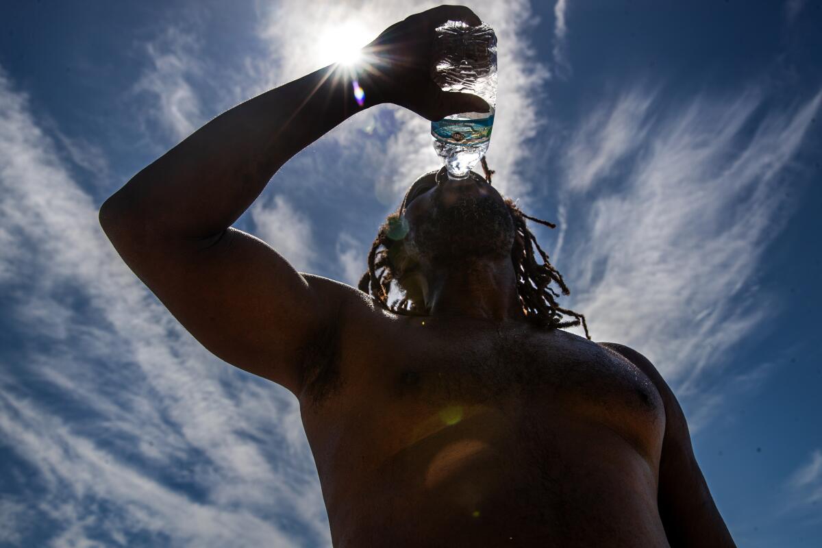 A shirtless man gulps water from a plastic bottle as the sun shines overhead.