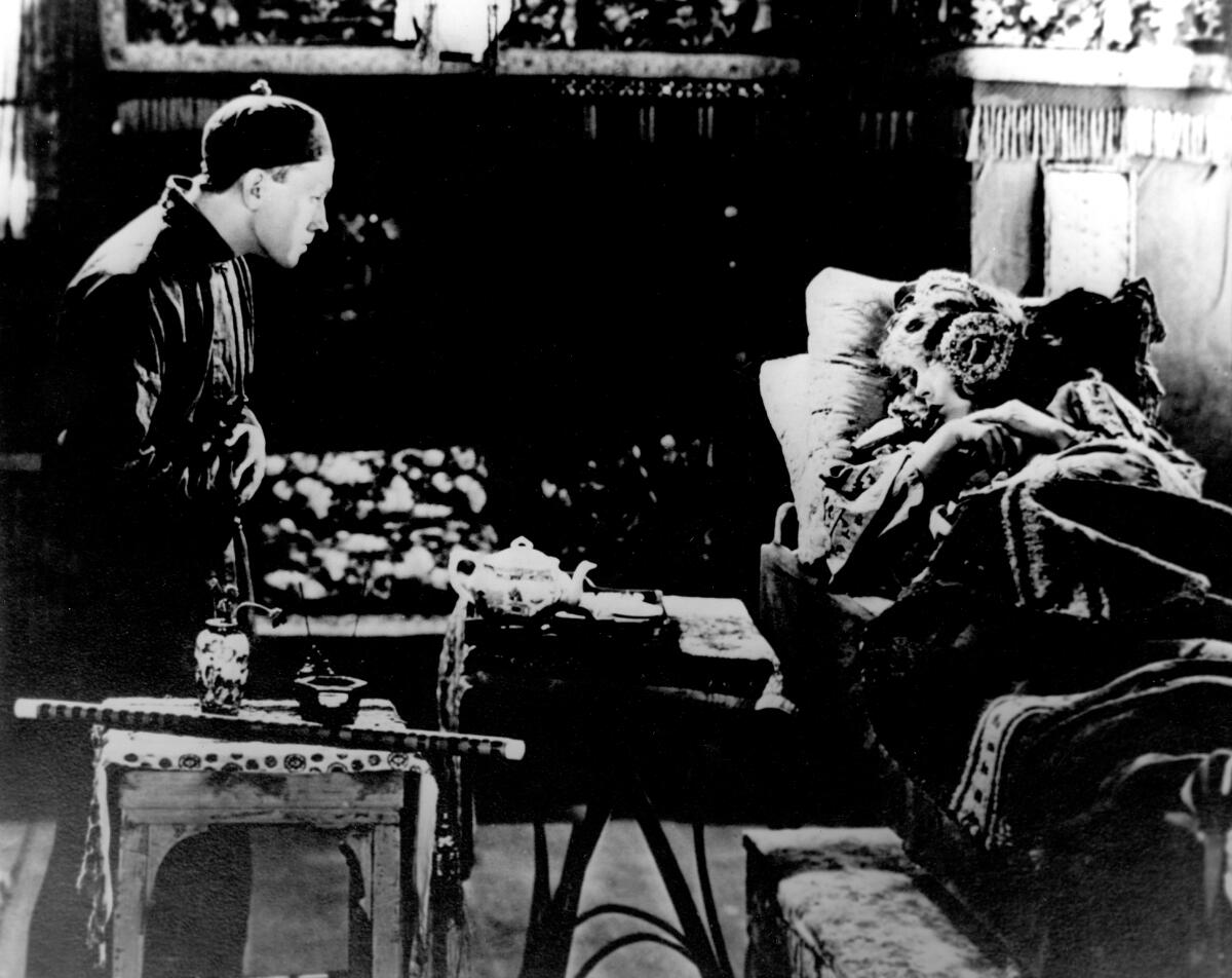 En una escena de película en blanco y negro, un hombre sentado mira a una mujer acostada