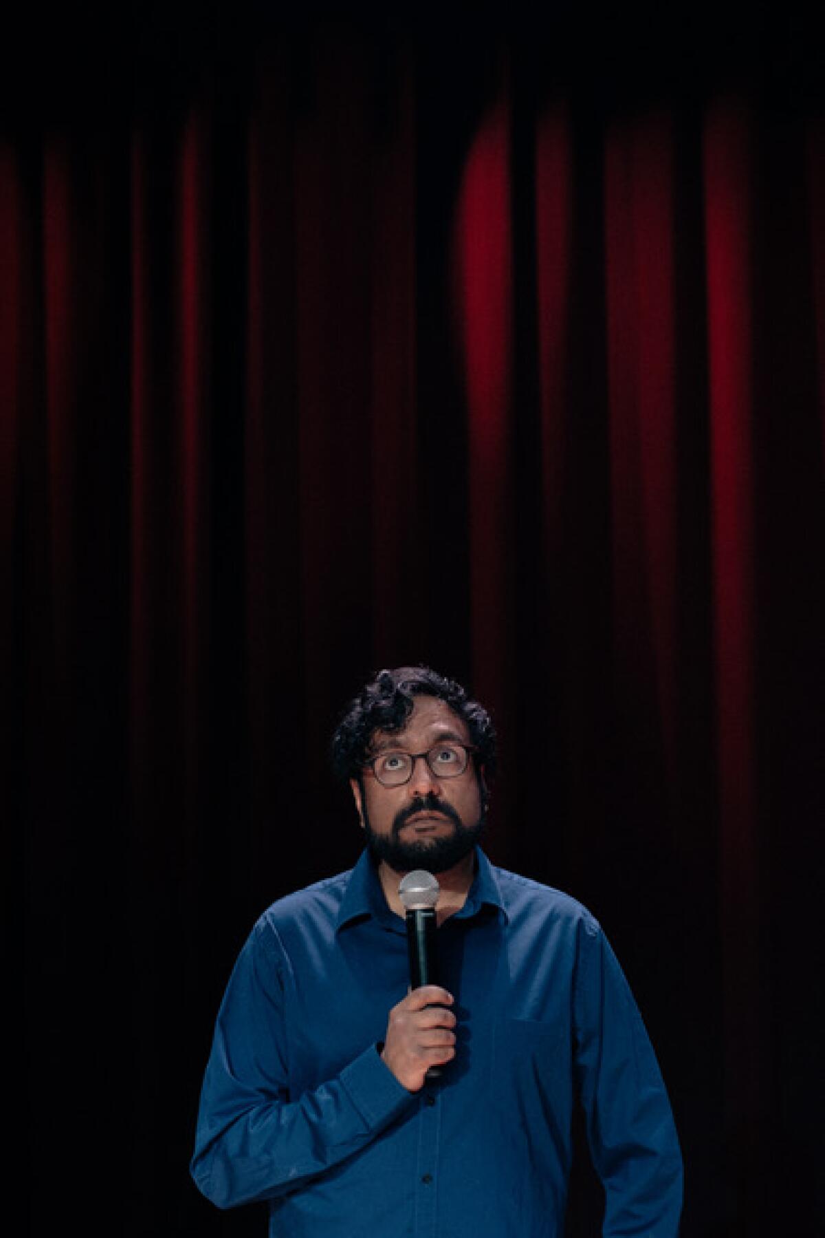 Hari Kondabolu holding a microphone in front of a red curtain