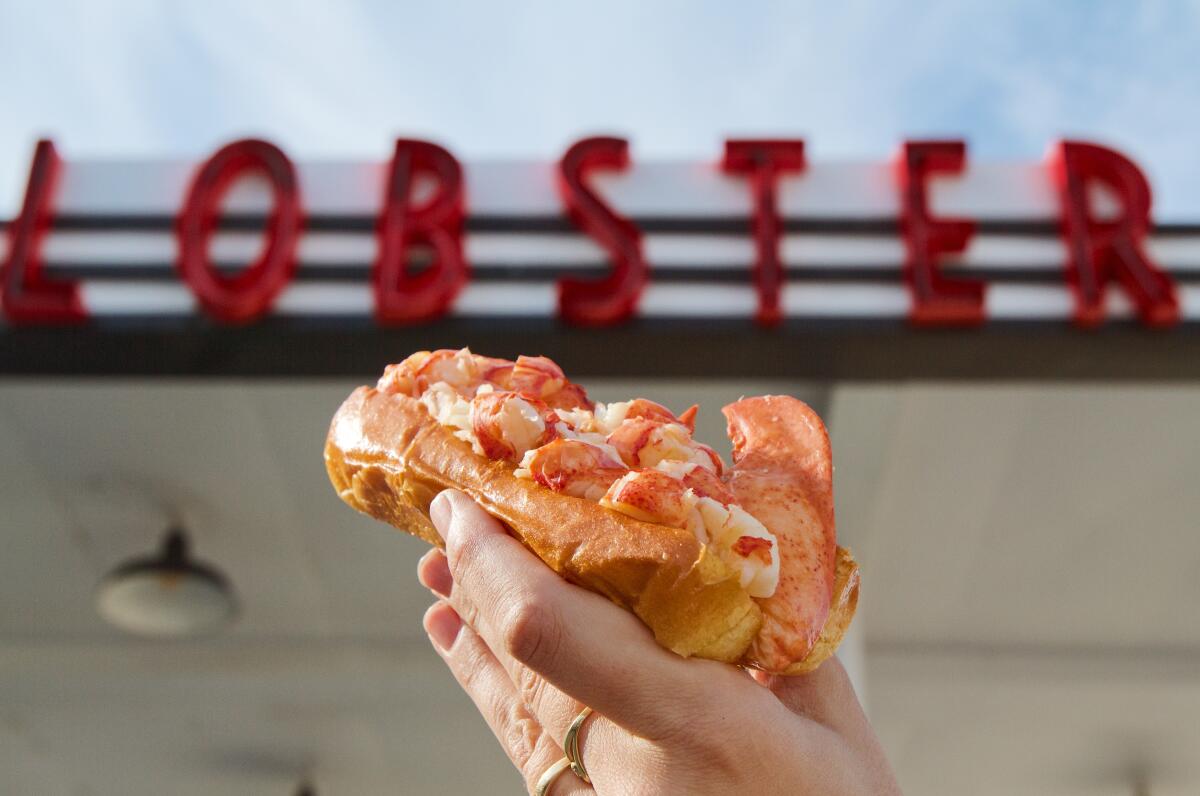 A hand holds a massive lobster roll in front of a vintage gas station's red "LOBSTER" signage.