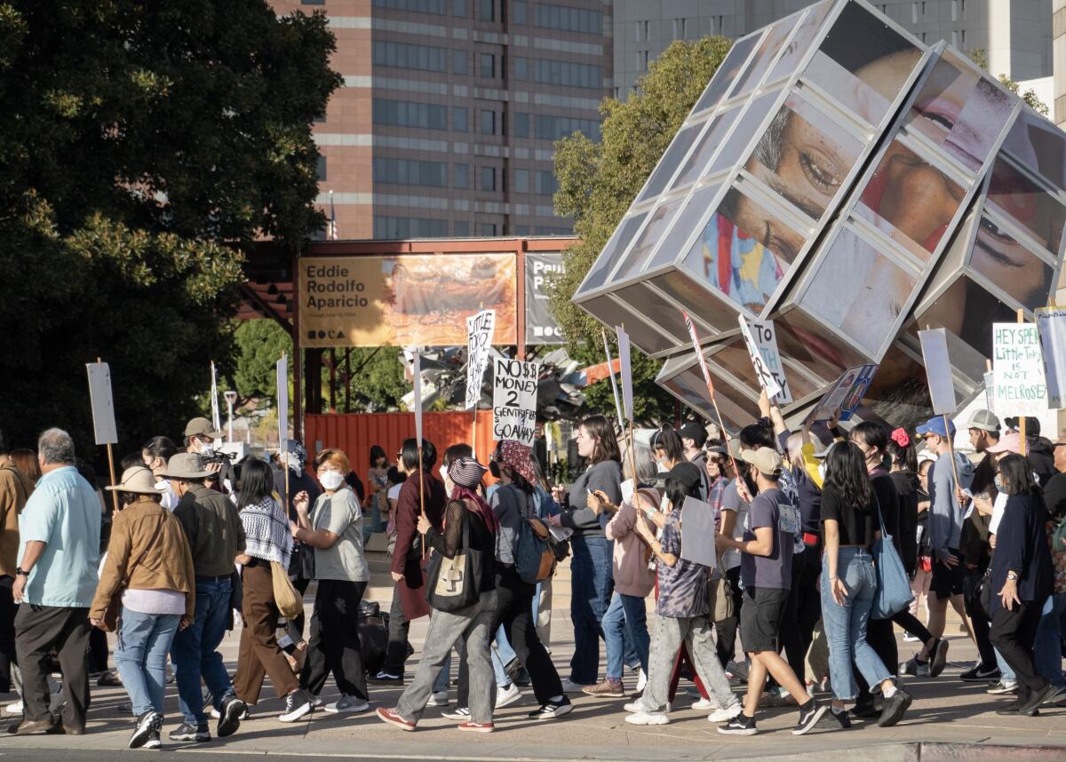 People holding signs walk past a big cube-like sculpture.