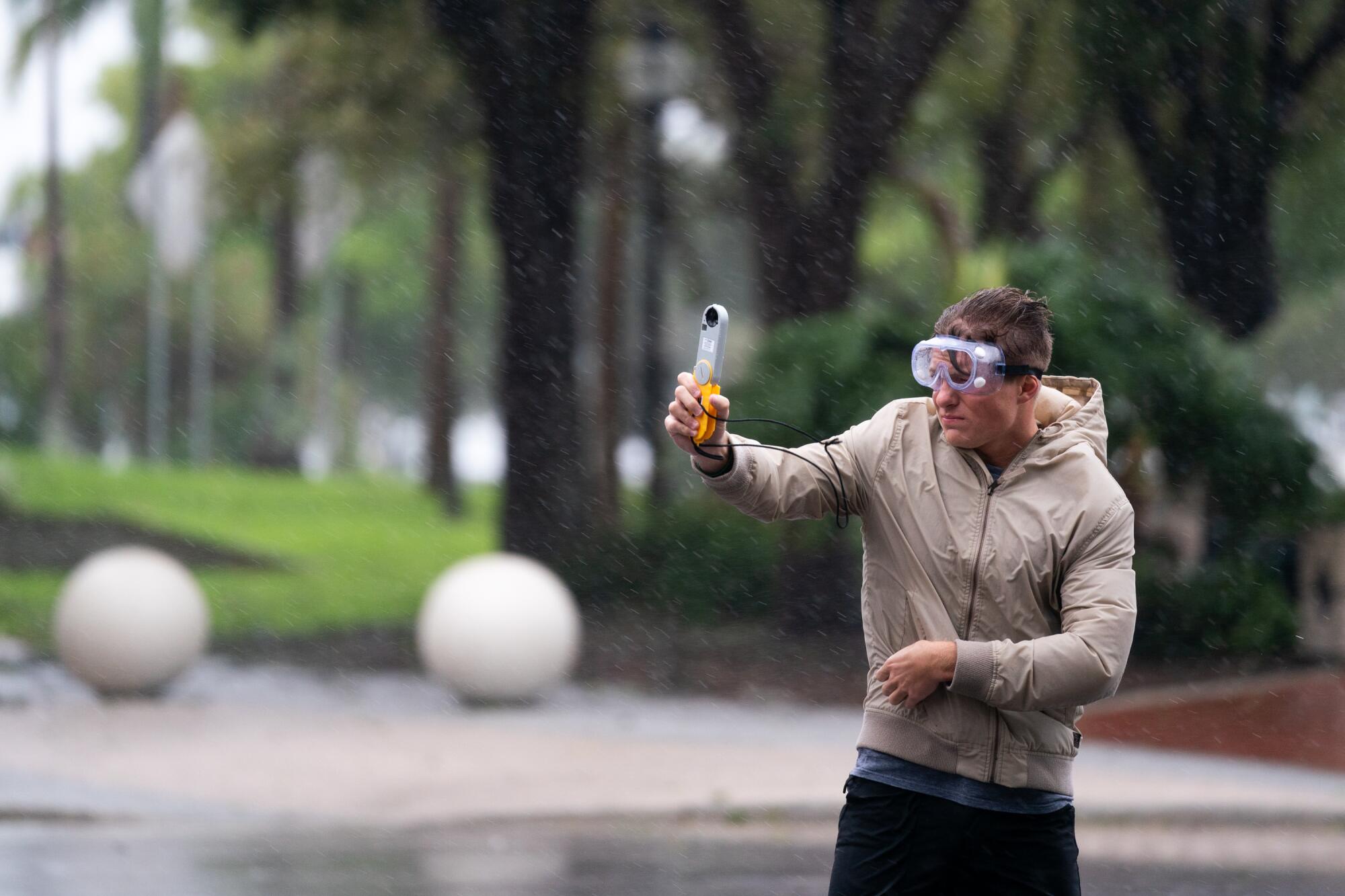 A man in goggles with a hand-held instrument stands in the wind and rain.