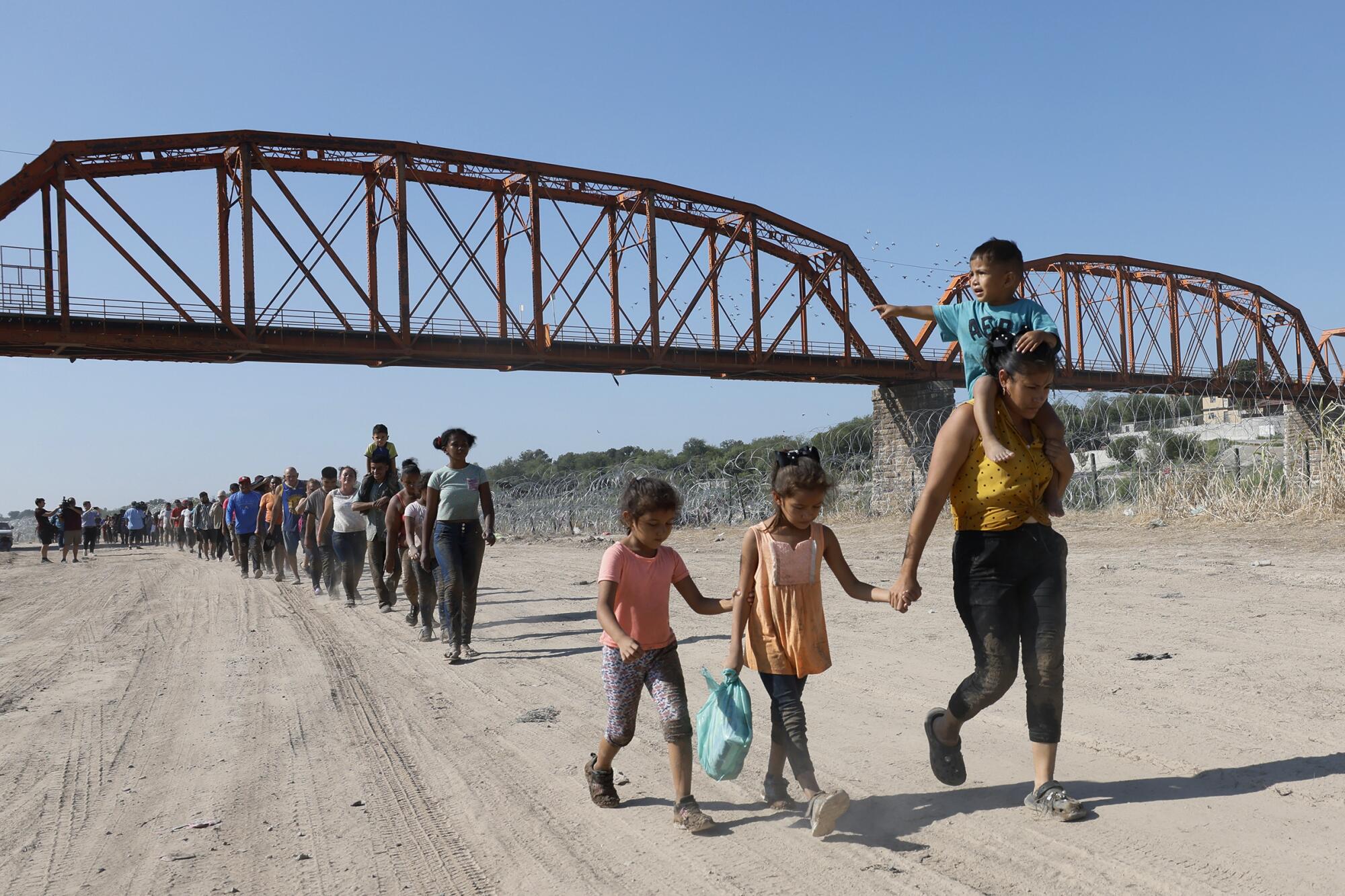 Hundreds of migrants walk in a long line on a sandy track under a bridge