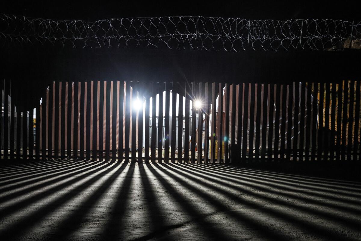 Lights shine through the vertical slats of a fence at night.