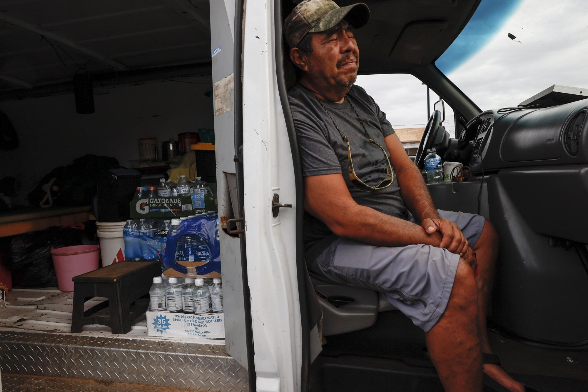 A man sits in the passenger seat of a van. In the back is bottled water, buckets and a stepstool.