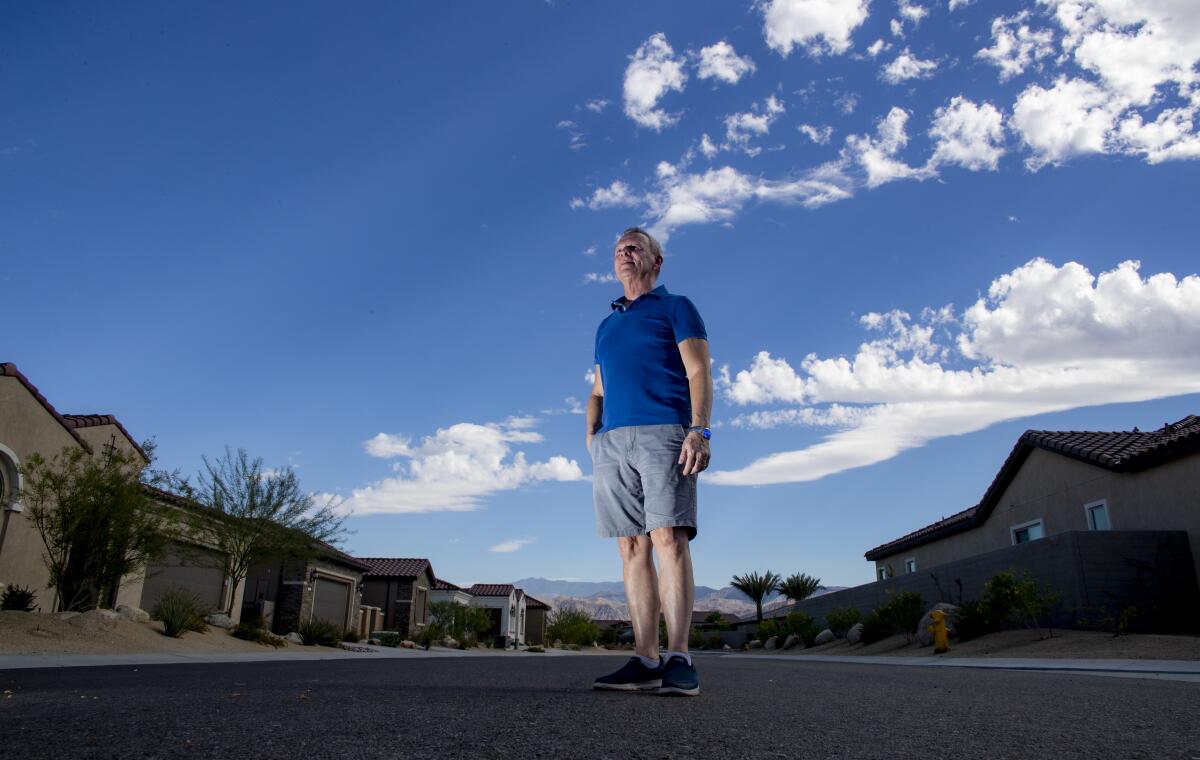 A man in a blue shirt and gray shorts stands on a street, flanked by homes, with blue skies as a backdrop