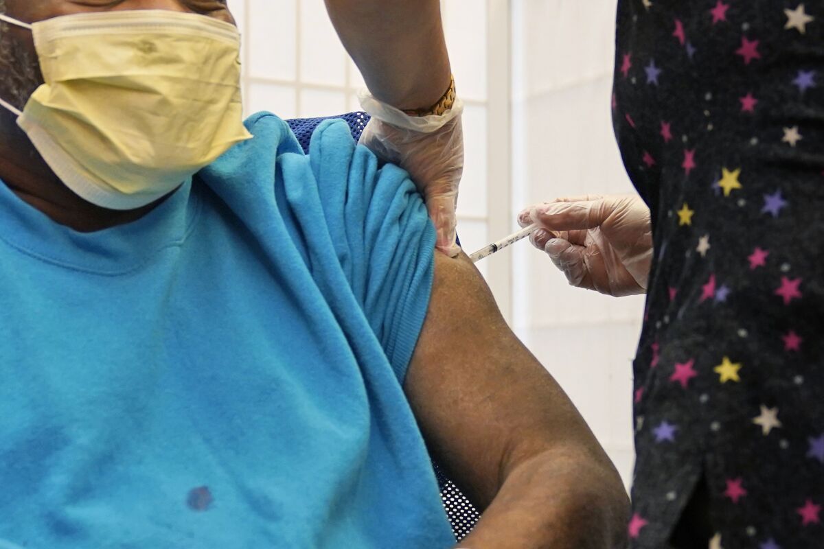 A pair of gloved hands administer a shot into the arm of a masked person in a blue shirt