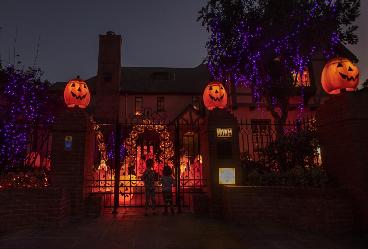 Jack-o'-lanterns glow atop pillars of a gated home, where two children stand looking at more pumpkins behind the gates 